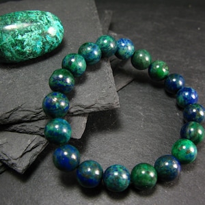 May include: A green and blue gemstone bracelet made of round beads. The bracelet is on a black slate surface. There is a large green and blue gemstone next to the bracelet.