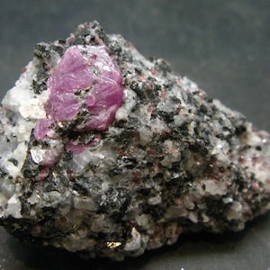 May include: A large, pink ruby crystal embedded in a dark gray rock matrix. The ruby is surrounded by smaller, white crystals.