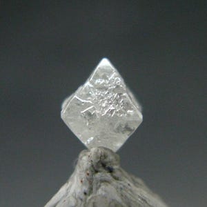 Pode incluir: Um cristal transparente e translúcido em forma de diamante com facetas angulares nítidas e uma superfície cristalina texturizada. A pedra apresenta uma estrutura geométrica semelhante a um octaedro com um topo pontiagudo e arestas.