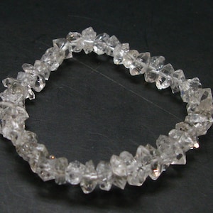 Puede incluir: Un brazalete de cuarzo transparente compuesto por cristales de cuarzo facetados de forma irregular. Los cristales están ensartados para formar una forma circular. El brazalete se muestra sobre un fondo oscuro.