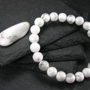 May include: A white howlite gemstone bracelet with white howlite beads. The bracelet is on a black slate surface, and there is a white howlite stone next to it.