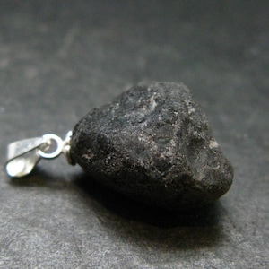 Puede incluir: Un colgante de piedra en bruto de color gris carbón oscuro con una textura rugosa e irregular. La piedra está unida a una anilla de metal plateado pulido, que cuenta con un pequeño aro circular para colgar la pieza en una cadena.