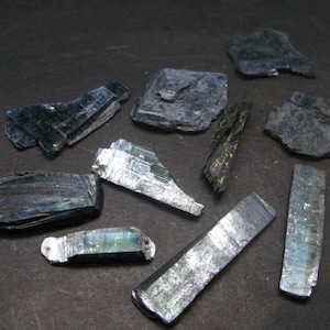 Könnte beinhalten: Eine Sammlung roher, ungeschliffener Kyanitkristalle in Blau-, Grün- und Silbertönen. Die Kristalle variieren in Größe und Form, einige erscheinen als flache, geschichtete Stücke, andere als längliche, prismatische Formen. Die Kristalle sind auf einer dunklen Oberfläche angeordnet.