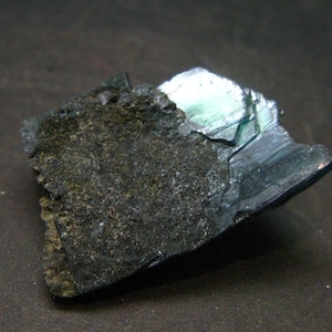 Könnte beinhalten: Dunkles, strukturiertes Mineralexemplar mit einer rauen, dunkelgrauen Außenseite und einem Bereich mit schillernder, türkisblauer Kristallstruktur. Das Mineral hat ein geschichtetes Aussehen und liegt vor einem dunklen Hintergrund.