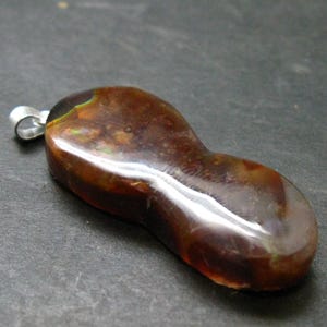 May include: A polished, peanut-shaped pendant featuring deep brown and amber tones with subtle iridescent green flashes throughout the stone. The pendant is attached to a simple silver-toned metal bail at the top, showcasing a smooth, glossy surface.