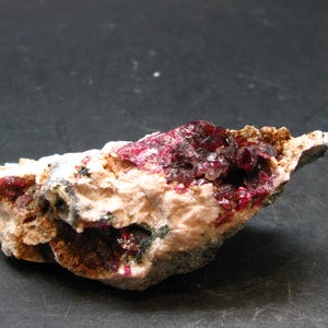 Puede incluir: Un espécimen mineral natural con un grupo de cristales rojo oscuro incrustados en una matriz de roca beige y blanca. Los cristales tienen una apariencia brillante, similar a una gema, que contrasta con la textura rugosa de la piedra. La composición es de tonos terrosos.