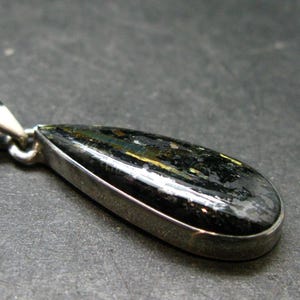 May include: A teardrop-shaped pendant featuring a dark, polished stone with visible streaks of gold and silver metallic inclusions, encased in a smooth silver-toned metal bezel setting with a matching silver-toned triangular bail at the top.