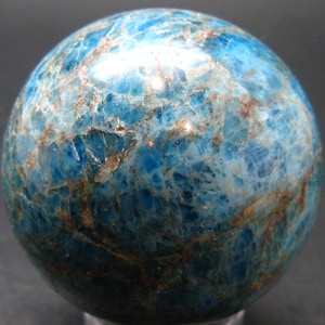 May include: A polished sphere of blue apatite, a gemstone with a marbled appearance. The stone's surface features a mix of light and dark blue hues, with streaks of brown and tan. The sphere is a decorative item.