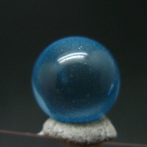 Puede incluir: Una canica de cristal azul, lisa y esférica, con un aspecto translúcido. El objeto presenta una superficie pulida que refleja la luz, creando un brillo intenso en su curva superior. Se apoya sobre una pequeña base de color claro.