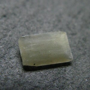 May include: A small, rectangular, translucent gemstone with a pale, muted green hue. The stone features a smooth, polished surface with subtle internal variations and a flat, geometric shape, showcasing a soft, light-reflecting quality throughout.