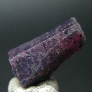 May include: A deep purple, faceted, rough-cut gemstone with a pointed tip. The gemstone has a natural, unpolished look.
