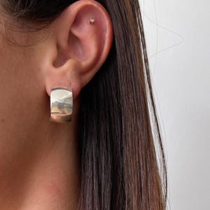 May include: A close-up of a silver-coloured, chunky hoop earring. The earring is a thick, rounded rectangle shape. The earring is worn on the ear of a person with long, dark brown hair. The background is a plain white wall.