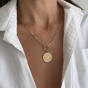 May include: A gold-toned necklace featuring a long, rectangular link chain and a circular pendant with an intricate, embossed floral pattern. The pendant is attached to the chain by a horizontal toggle bar closure that rests above the coin charm.
