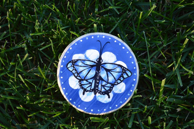 K&ouml;nnte beinhalten: Eine handbemalte Holzscheibe mit blauem Hintergrund und wei&szlig;en Punkten am Rand. Ein schwarzer Schmetterling mit blauen Fl&uuml;geln ist in der Mitte der Scheibe gemalt, mit einer wei&szlig;en Blume dahinter.