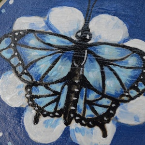 K&ouml;nnte beinhalten: Ein blauer Schmetterling mit schwarzen Markierungen ist auf eine runde Holzscheibe gemalt. Der Schmetterling befindet sich in der Mitte der Scheibe und ist von wei&szlig;en Bl&uuml;tenbl&auml;ttern umgeben. Der Hintergrund der Scheibe ist dunkelblau.
