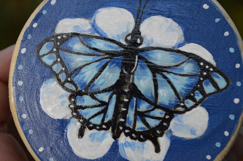 K&ouml;nnte beinhalten: Ein blauer Schmetterling mit schwarzen Markierungen ist auf eine runde Holzscheibe gemalt. Der Schmetterling befindet sich in der Mitte der Scheibe und ist von wei&szlig;en Bl&uuml;tenbl&auml;ttern umgeben. Der Hintergrund der Scheibe ist dunkelblau.
