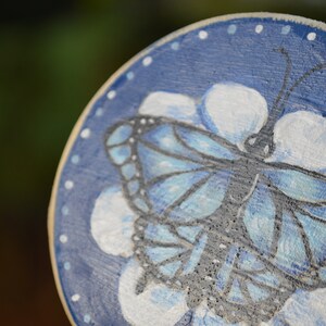 K&ouml;nnte beinhalten: Eine runde Holzscheibe mit blauem Hintergrund und wei&szlig;en Punkten. Auf der Scheibe ist ein blauer Schmetterling mit schwarzen Adern gemalt, umgeben von wei&szlig;en Bl&uuml;tenbl&auml;ttern.