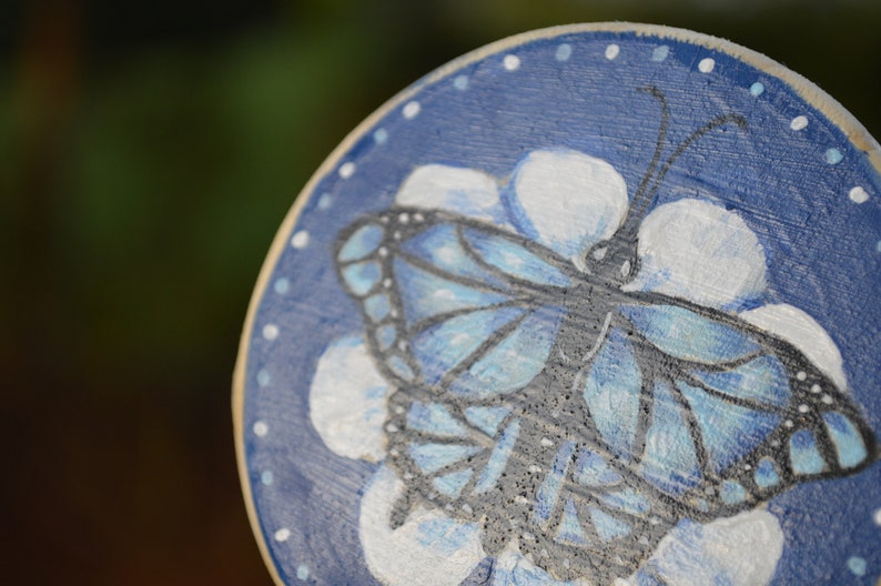 K&ouml;nnte beinhalten: Eine runde Holzscheibe mit blauem Hintergrund und wei&szlig;en Punkten. Auf der Scheibe ist ein blauer Schmetterling mit schwarzen Adern gemalt, umgeben von wei&szlig;en Bl&uuml;tenbl&auml;ttern.
