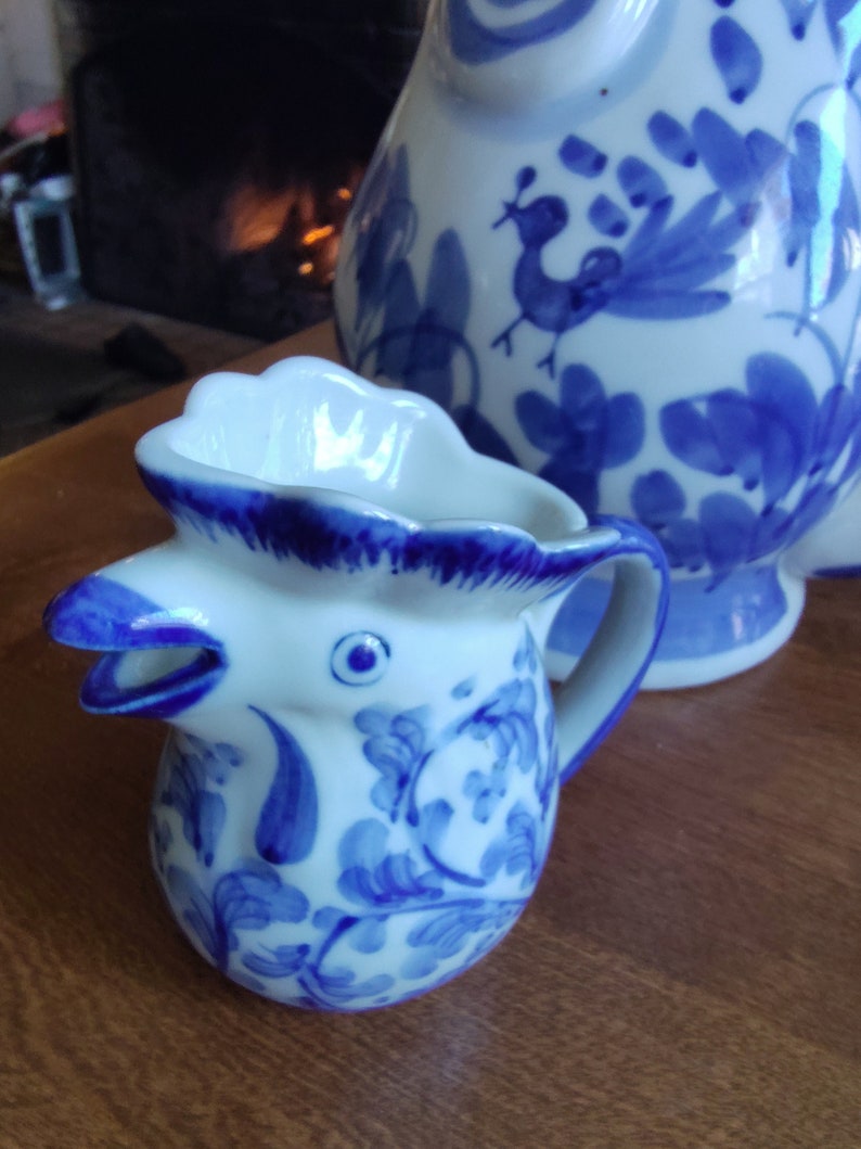 May include: A white and blue ceramic creamer in the shape of a rooster's head, with a matching pitcher in the background. The creamer has a handle and is decorated with blue floral patterns. The pitcher also features blue floral designs and a peacock.