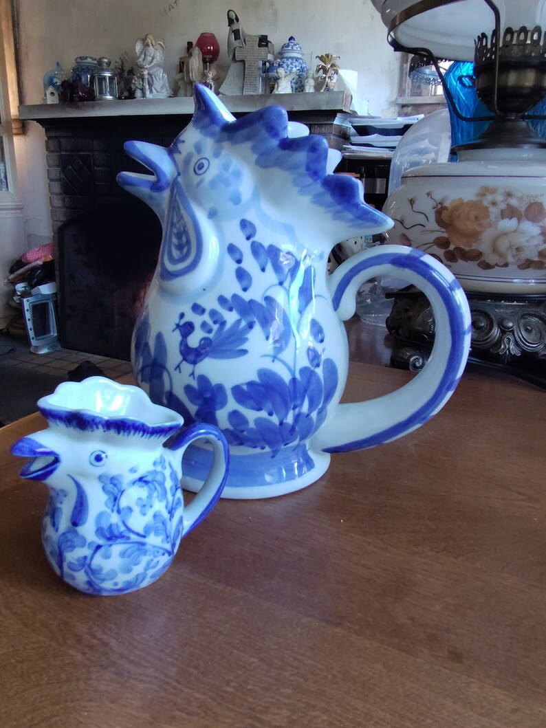 May include: Two ceramic pitchers with a rooster design. The larger pitcher is white with blue floral and rooster details, and a curved handle. The smaller pitcher has a similar design. Both are on a wooden surface.