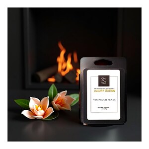 Puede incluir: Un paquete negro de ceras perfumadas con el texto "VIN PHUCH PEAKS" y "LUXURY EDITION". Dos decoraciones florales de color melocot&oacute;n con hojas verdes est&aacute;n delante del paquete. El fondo muestra una chimenea con fuego encendido.