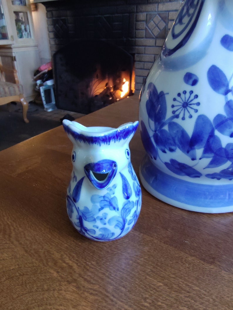 May include: A small, white and blue ceramic creamer in the shape of a bird, with a spout for pouring. The creamer is decorated with blue floral patterns. A larger matching pitcher is visible in the background. Both are on a wooden surface.