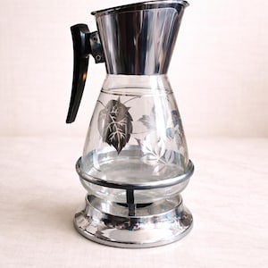 May include: Vintage glass and chrome pitcher with a black handle. The clear glass body features etched leaf designs. The pitcher sits on a chrome base and has a chrome top.