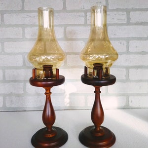 Two vintage amber glass pedestal lamps are shown side by side against a white brick background. Their warm honey-colored chimneys and turned wood bases are evenly matched, creating a symmetrical decorative pair.