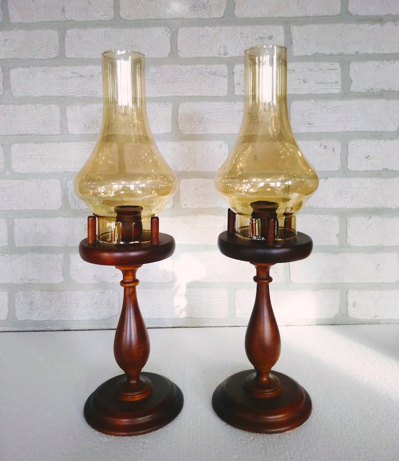 Two vintage amber glass pedestal lamps are shown side by side against a white brick background. Their warm honey-colored chimneys and turned wood bases are evenly matched, creating a symmetrical decorative pair.