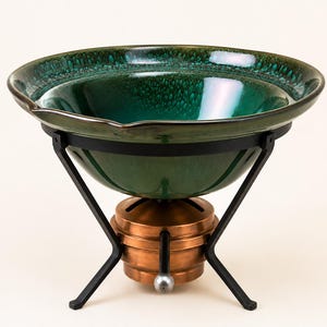 May include: A deep green ceramic bowl with a glossy, mottled glaze finish rests inside a black metal stand. The stand is supported by a central copper-colored cylindrical base featuring a small silver-toned knob on the front exterior.