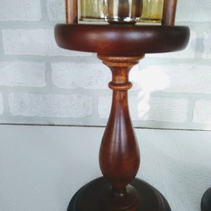 This image shows the wooden pedestal base with its elegant turned shape and polished medium-brown finish. The glass chimney sits securely above the base, supported by vertical wooden posts.