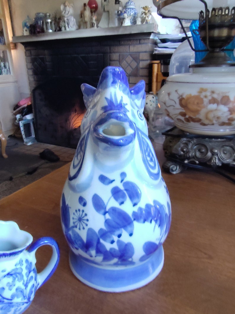 May include: A ceramic teapot with a blue and white floral design. The teapot is shaped like a pig with a spout and handle. The teapot is sitting on a wooden table. A small creamer with a matching design is in the foreground.