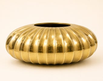 May include: A low, round, metallic gold bowl with a polished finish and a distinct vertical ribbed texture. The vessel features a wide, circular opening at the top and a flattened, disc-like shape that tapers toward the center opening.