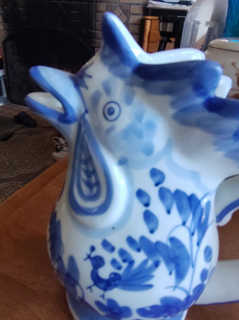 May include: A white ceramic rooster-shaped pitcher with blue hand-painted floral and peacock designs. The pitcher has a long spout and a handle. The rooster's head is tilted back, and its beak is open. The pitcher is on a wooden surface.