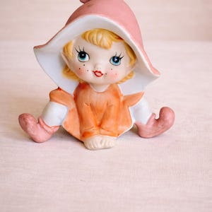 May include: A ceramic figurine of a seated elf with blonde hair, wearing a large pink pointed hat, an orange tunic, and white pants. The elf has blue eyes, red lips, and freckles, with pink curled-toe shoes extending from the base of the figure.