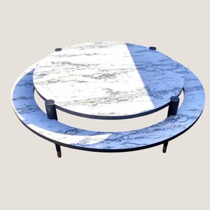 May include: A circular coffee table featuring a white faux marble top with grey veining, supported by a black metal frame. The design includes a lower circular shelf that follows the perimeter of the table, connected by vertical black metal supports.