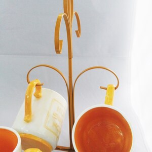 May include: A gold metal mug rack with four white ceramic mugs with yellow accents. The mugs are hanging upside down on the rack.