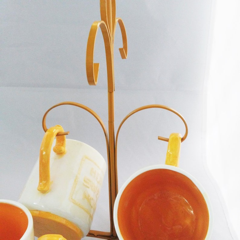 May include: A gold metal mug rack with four white ceramic mugs with yellow accents. The mugs are hanging upside down on the rack.