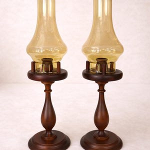 Two vintage amber glass pedestal lamps are shown side by side against a white brick background. Their warm honey-colored chimneys and turned wood bases are evenly matched, creating a symmetrical decorative pair.