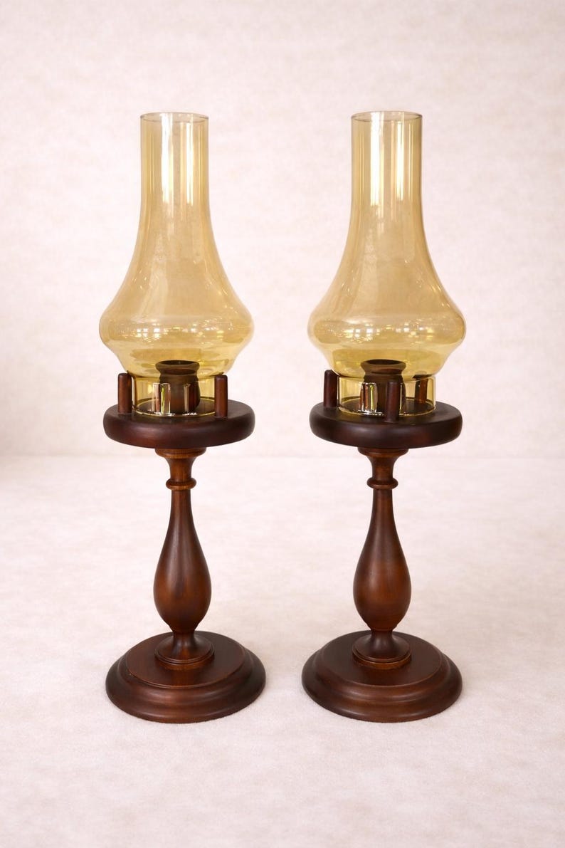Two vintage amber glass pedestal lamps are shown side by side against a white brick background. Their warm honey-colored chimneys and turned wood bases are evenly matched, creating a symmetrical decorative pair.