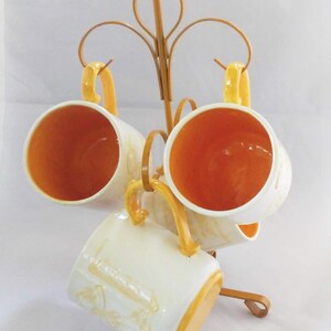 May include: Set of four white ceramic mugs with yellow accents and a floral design. The mugs are hanging on a gold metal stand.