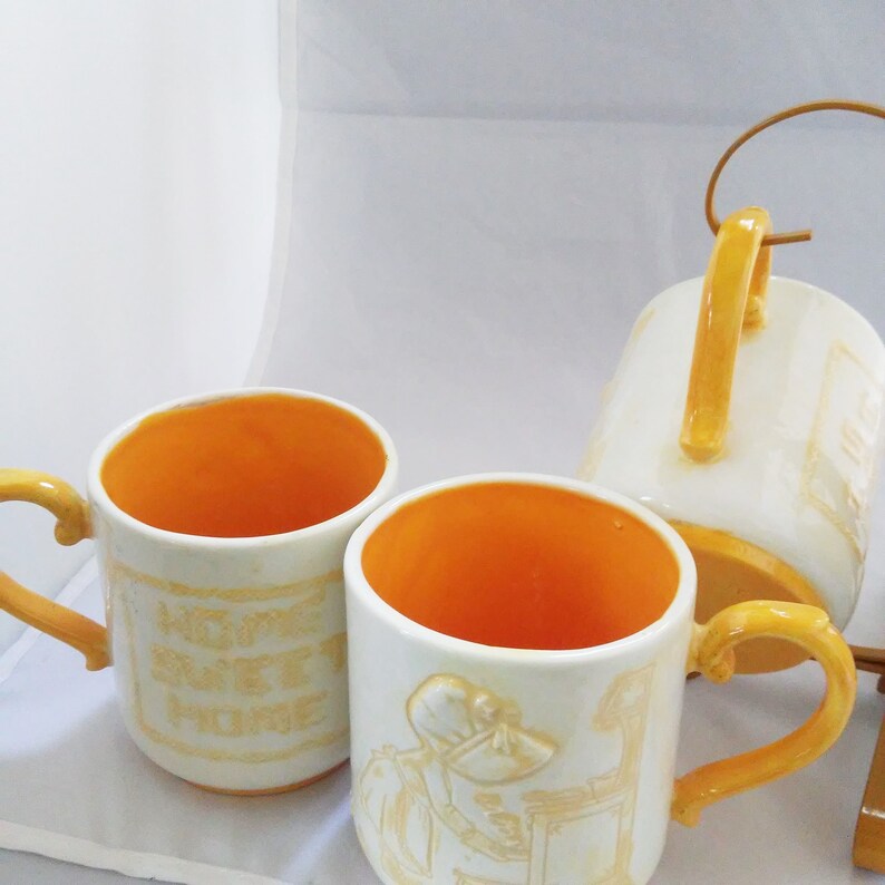May include: Two yellow and white ceramic mugs with a yellow handle. One mug has the text "Home Sweet Home" on it. The other mug has a picture of a woman in a bonnet and a dress.