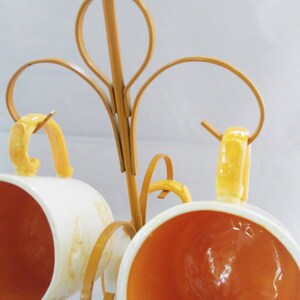 May include: A gold metal mug rack with three white ceramic mugs with yellow accents. The mugs are hanging from the rack by their handles.