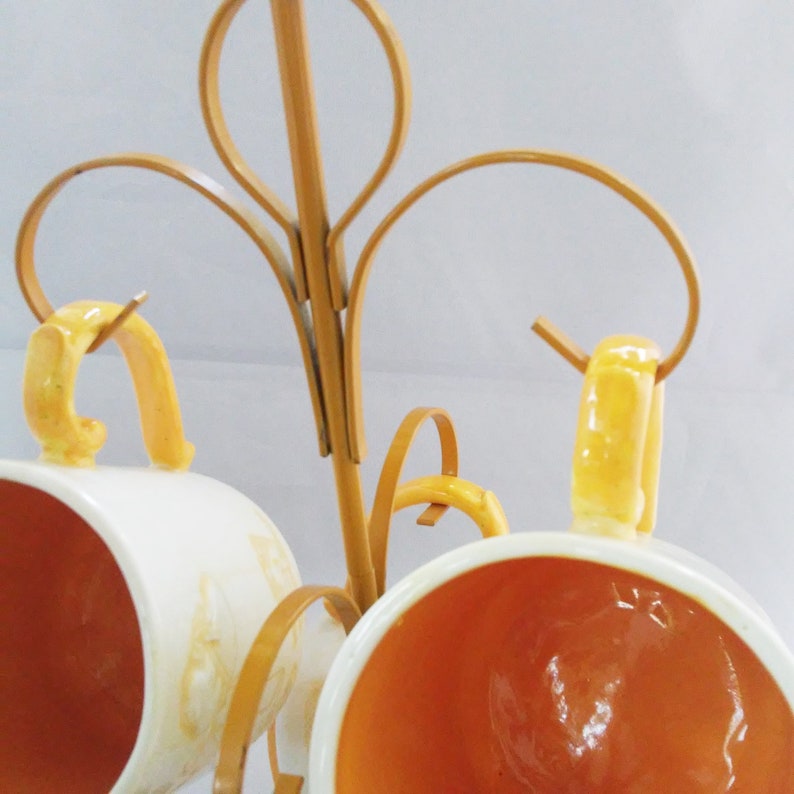 May include: A gold metal mug rack with three white ceramic mugs with yellow accents. The mugs are hanging from the rack by their handles.