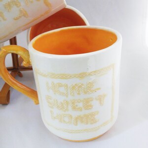 May include: White ceramic mug with a yellow rim and handle. The mug has a yellow embroidered design that says "Home Sweet Home".