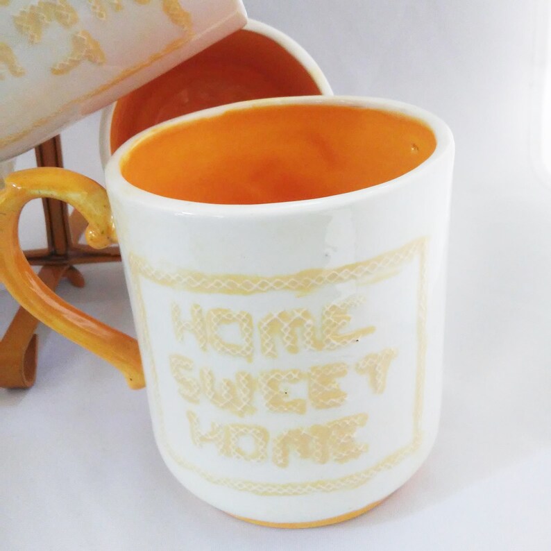 May include: White ceramic mug with a yellow rim and handle. The mug has a yellow embroidered design that says "Home Sweet Home".