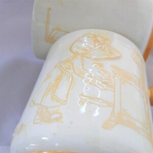 May include: A white ceramic mug with a yellow design of a girl in a dress and hat. The girl is standing next to a table with a drawer. The mug has a gold rim.