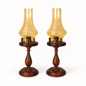Two vintage amber glass pedestal lamps are shown side by side against a white brick background. Their warm honey-colored chimneys and turned wood bases are evenly matched, creating a symmetrical decorative pair.