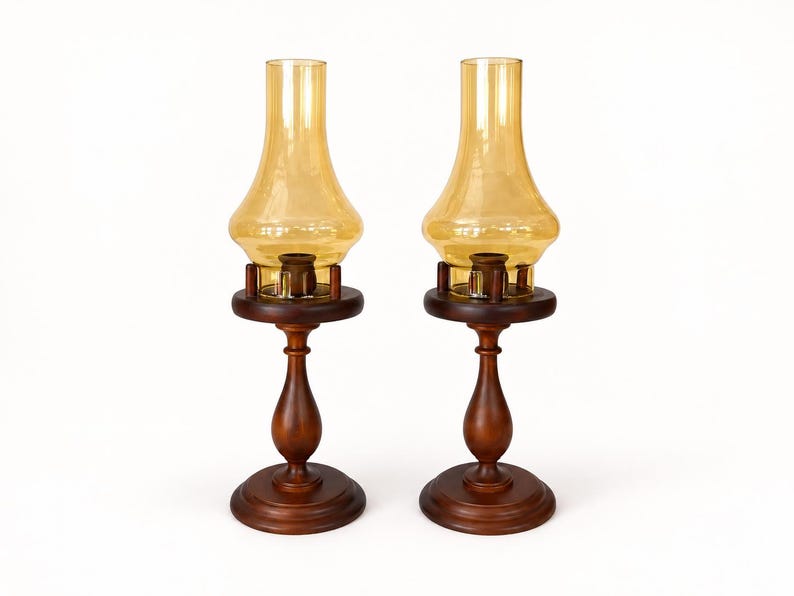 Two vintage amber glass pedestal lamps are shown side by side against a white brick background. Their warm honey-colored chimneys and turned wood bases are evenly matched, creating a symmetrical decorative pair.