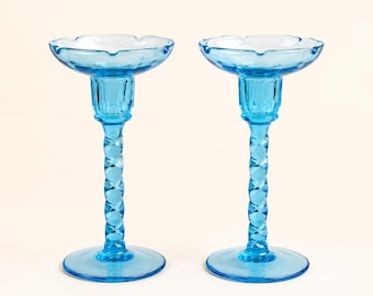 May include: A pair of matching bright blue glass candle holders featuring a wide, scalloped rim, a cylindrical neck, and a central stem with a distinct twisted, spiral pattern. Each piece stands on a circular base, showcasing a translucent finish.